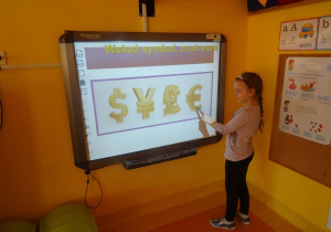 Dziewczynka stoi pod tablicą interaktywną i wskazuje znak euro.